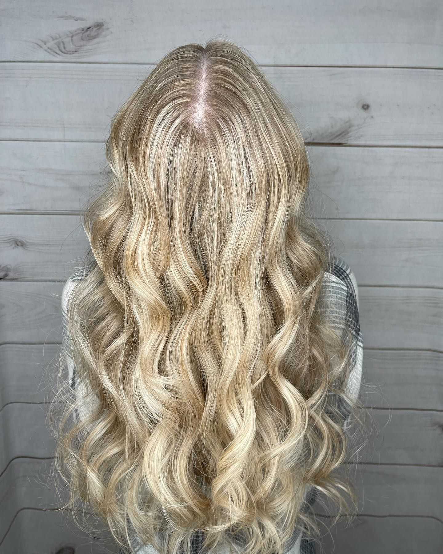 Long wavy blonde hair styled in loose curls, seen from the back against a wooden panel background.
