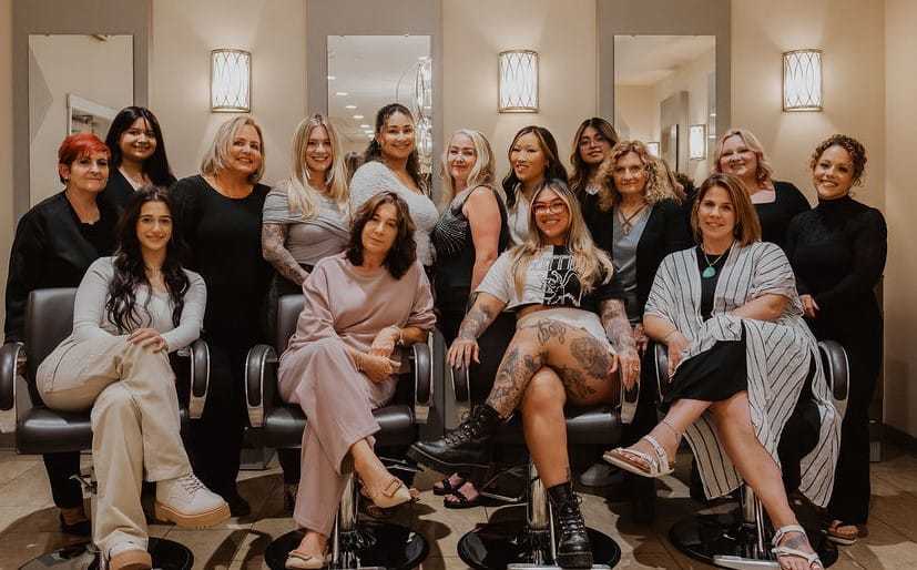 Group of fifteen women posing together in a salon, some seated, others standing behind them.