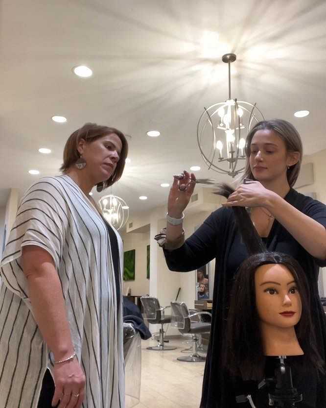 Hairdresser demonstrating haircut techniques with a mannequin in a salon, while a client observes.