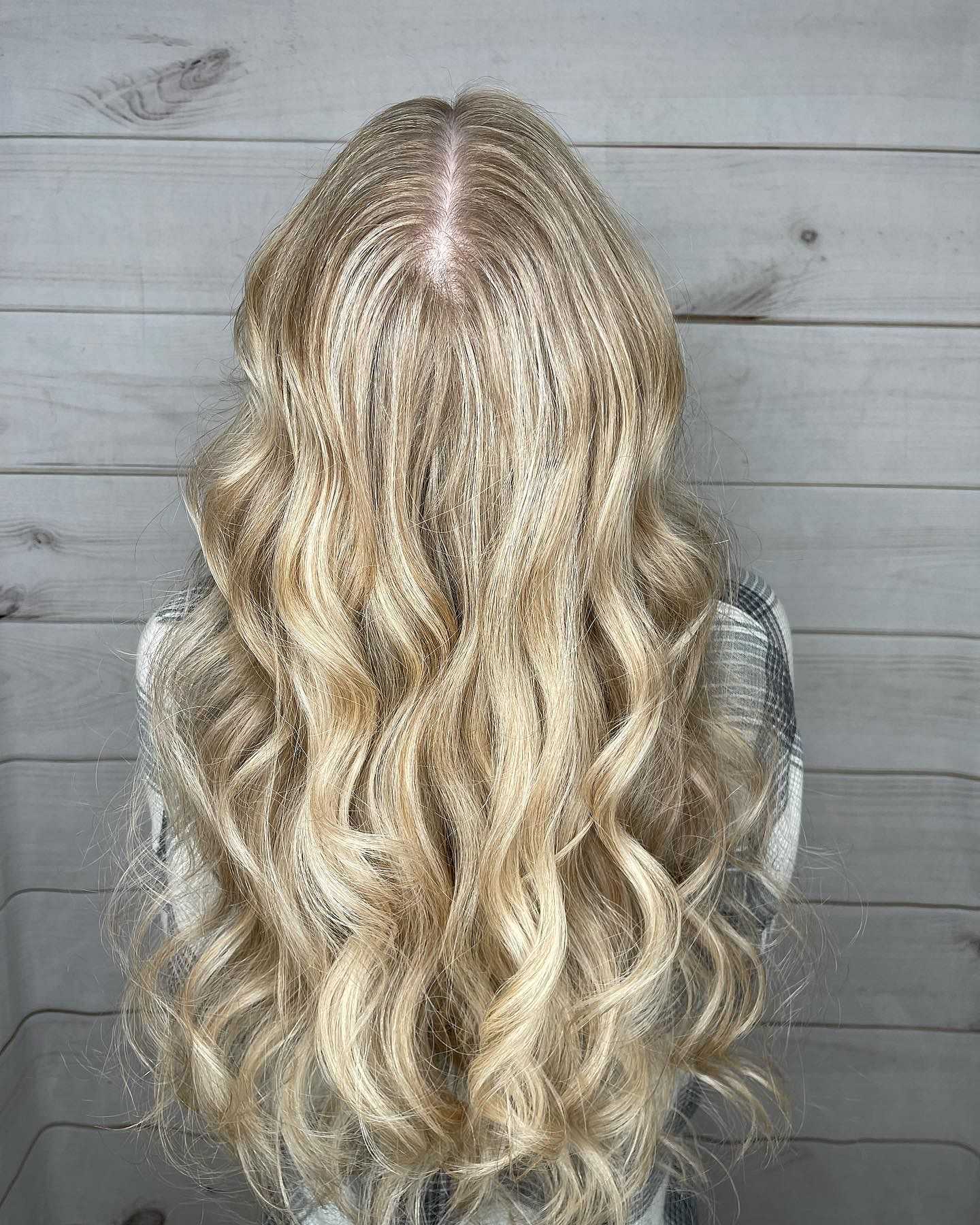 Long wavy blonde hair styled in loose curls, seen from the back against a wooden panel background.