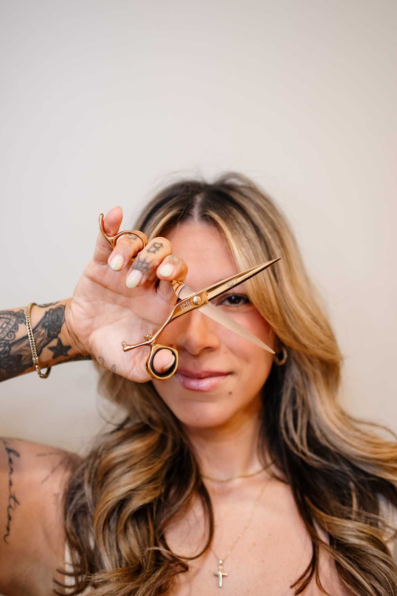 Hair stylist holding scissors, showcasing tattoos and long wavy hair.